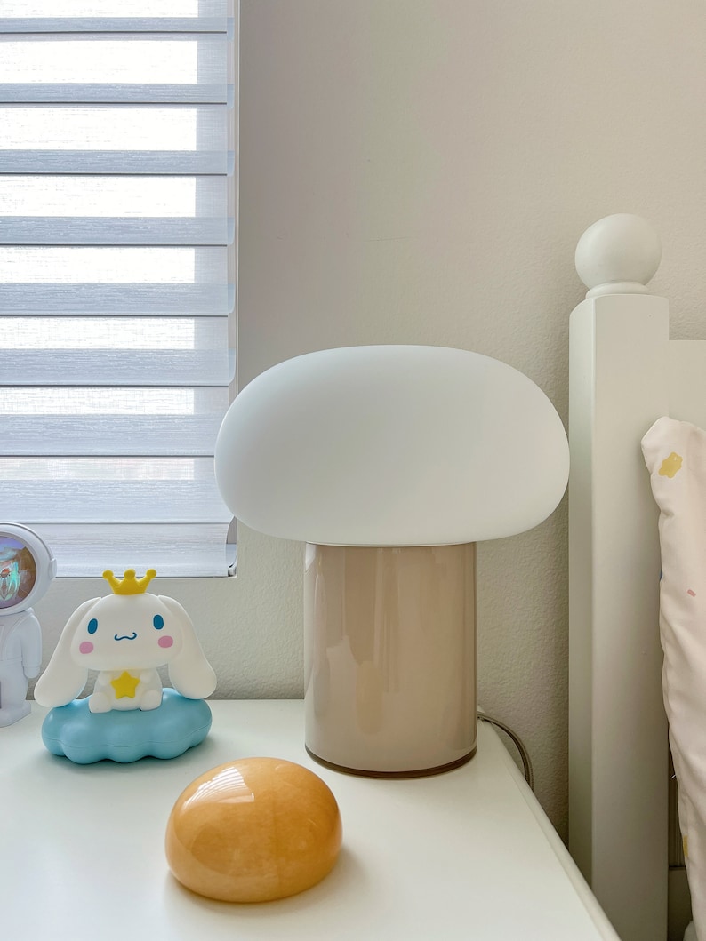 May include: A mushroom-shaped table lamp with a white shade and a beige base sits on a white nightstand. A cartoon character figurine and a small, orange, rounded object are also on the nightstand. The background includes a window with blinds and a white bed frame.