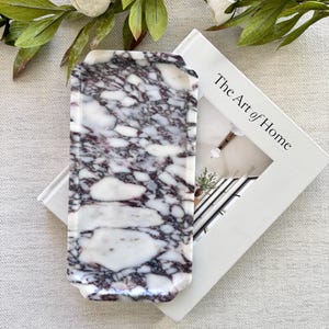 Puede incluir: Una bandeja rectangular de mármol con un patrón veteado blanco y morado. La bandeja está sobre un libro blanco titulado "The Art of Home". Flores blancas y hojas verdes en el fondo.