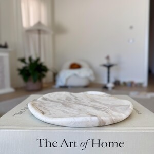 May include: A round, white and beige marble tray with a smooth, wavy edge resting on top of a light gray hardcover book titled The Art of Home. The tray features natural stone veining patterns in shades of cream, tan, and white across its surface.