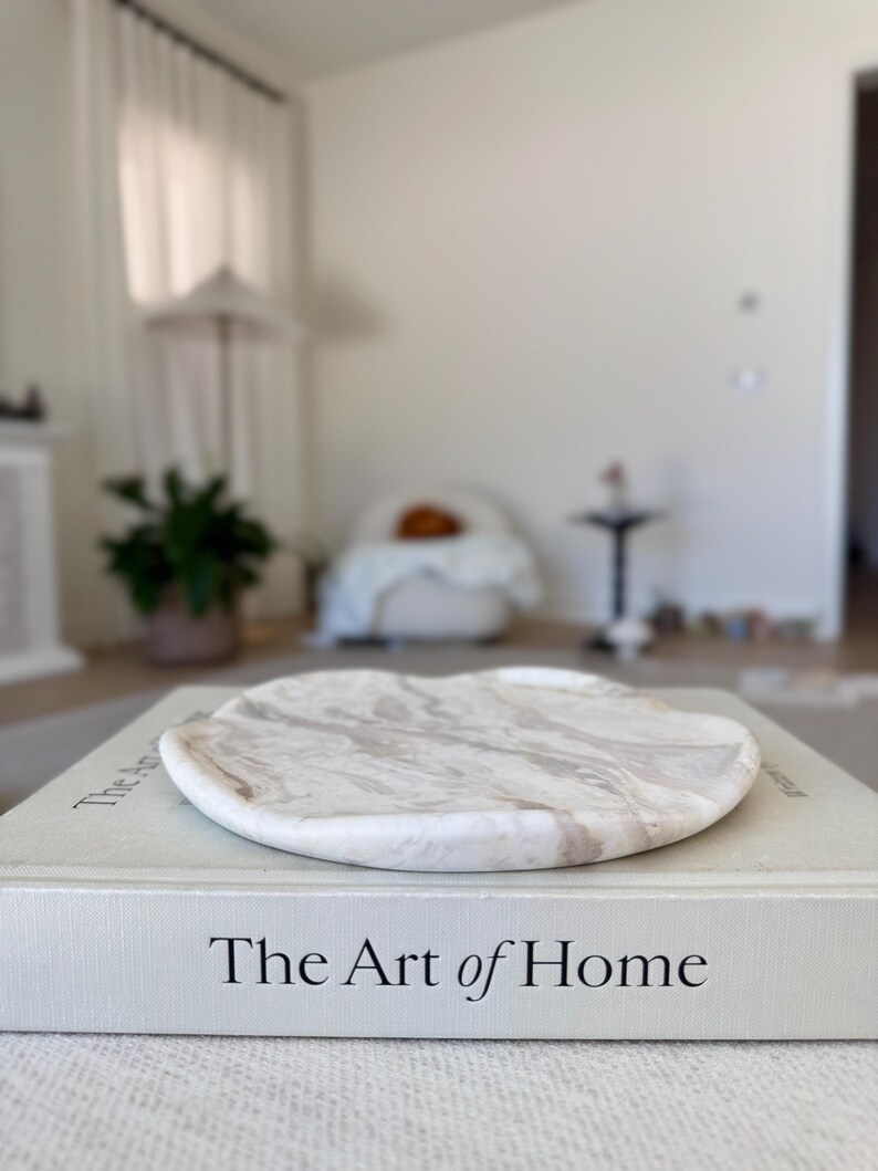 May include: A round, white and beige marble tray with a smooth, wavy edge resting on top of a light gray hardcover book titled The Art of Home. The tray features natural stone veining patterns in shades of cream, tan, and white across its surface.