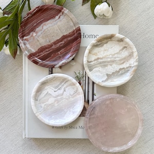 May include: Four round stone coasters in shades of red, white, and pink. The coasters feature unique veining patterns. One coaster is rose quartz. The coasters are on a book that says "Home".