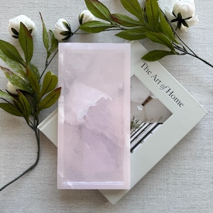 May include: A rectangular light pink tray with a smooth, polished finish and subtle white marbled veining. The tray is positioned on top of a book titled The Art of Home, which is decorated with a sprig of white flowers and green leaves.