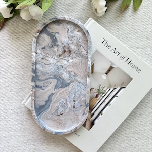 Puede incluir: Una bandeja decorativa ovalada con un diseño veteado en tonos grises, azules y beige. La bandeja descansa sobre un libro titulado "The Art of Home". Flores blancas y hojas verdes en el fondo.