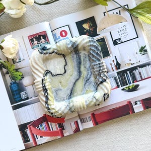 Peut inclure: Un plat carré en céramique texturée présentant un motif marbré dans des tons crème, jaune pâle et gris anthracite. La surface présente un bord ondulé et une finition brillante, avec des lignes organiques sombres tourbillonnant sur la base claire.