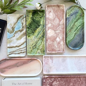 May include: A collection of seven rectangular and oval stone trays featuring various marble patterns in shades of green, pink, and white. The trays are arranged alongside a book titled The Art of Home and several white floral stems.
