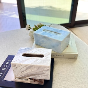 May include: Two rectangular tissue box covers made of polished stone. One cover features a light blue and white marbled pattern, while the other displays a grey and white swirling marble design. Each box has a centered oval opening for tissues.