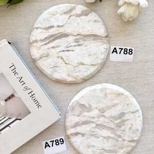 May include: Two circular stone coasters featuring a white and beige marbled pattern with natural veining. Each coaster is labeled with a small white tag, one marked A788 and the other marked A789, positioned near a book titled The Art of Home.