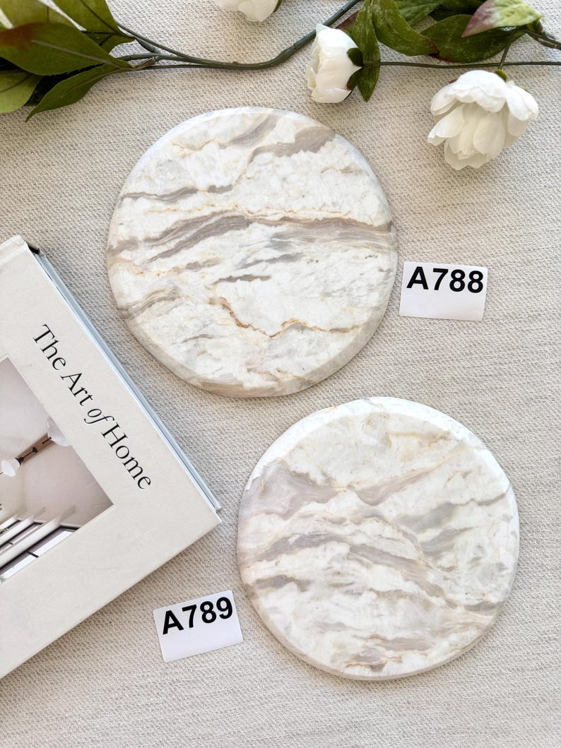 May include: Two circular stone coasters featuring a white and beige marbled pattern with natural veining. Each coaster is labeled with a small white tag, one marked A788 and the other marked A789, positioned near a book titled The Art of Home.