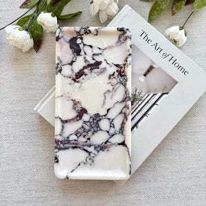 May include: A rectangular tray with a marble design in shades of white, burgundy, and black. The tray is resting on a book titled "The Art of Home" and is surrounded by white flowers and green leaves.