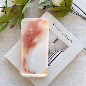 Puede incluir: Un jabonera rectangular translúcida con un diseño veteado en tonos blanco, melocotón y dorado. La jabonera está sobre un libro titulado "The Art of Home" con una ramita de flores blancas y hojas verdes.
