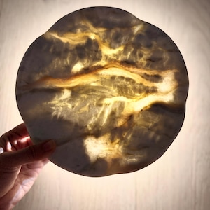 May include: A circular, translucent stone disc with an irregular, wavy edge. The surface features a marbled pattern of creamy white and deep amber veins, which glow with a warm, golden light when illuminated from behind, highlighting the natural texture.