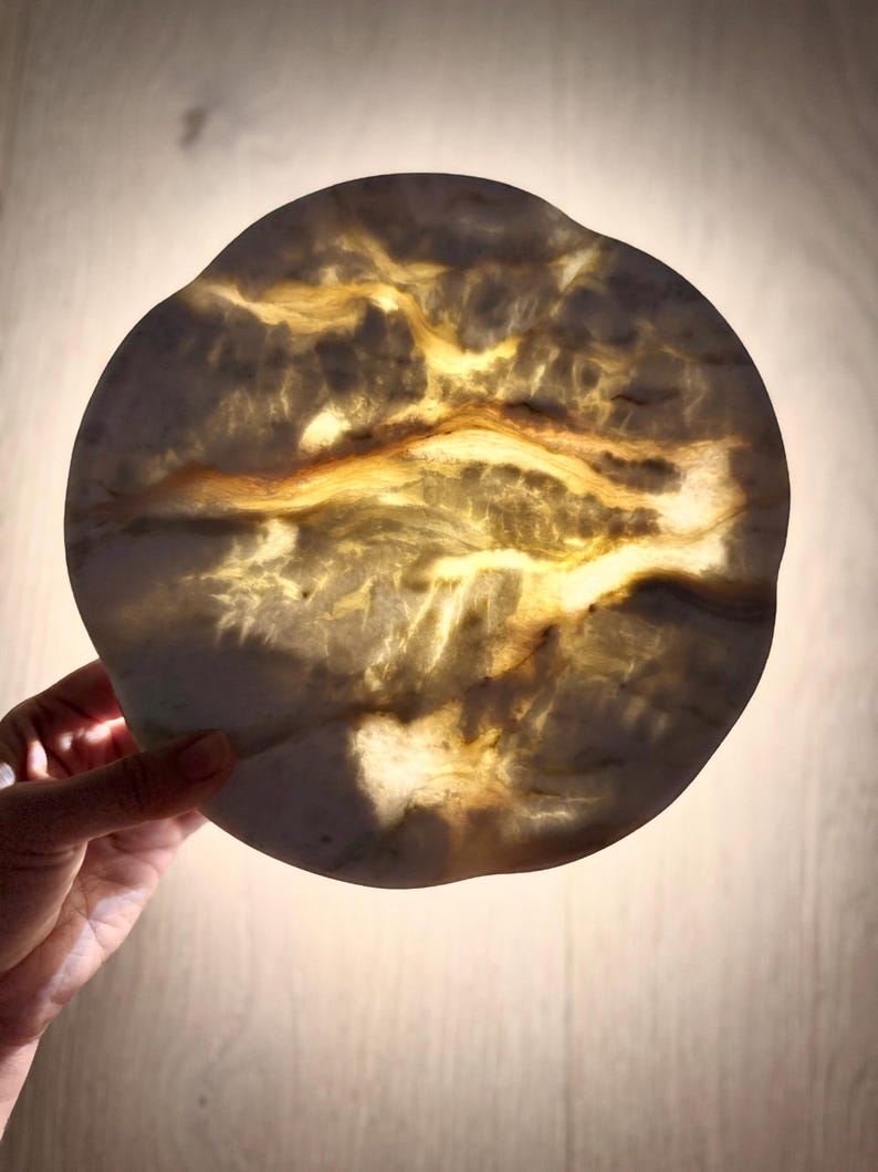 May include: A circular, translucent stone disc with an irregular, wavy edge. The surface features a marbled pattern of creamy white and deep amber veins, which glow with a warm, golden light when illuminated from behind, highlighting the natural texture.