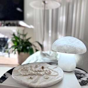 May include: A white marble tray holding a collection of pearl necklaces and a gold ring with a dark stone, placed on a book titled Sacred Spaces by Carley Summers, next to a glowing white mushroom-shaped lamp with a textured, stone-like surface.