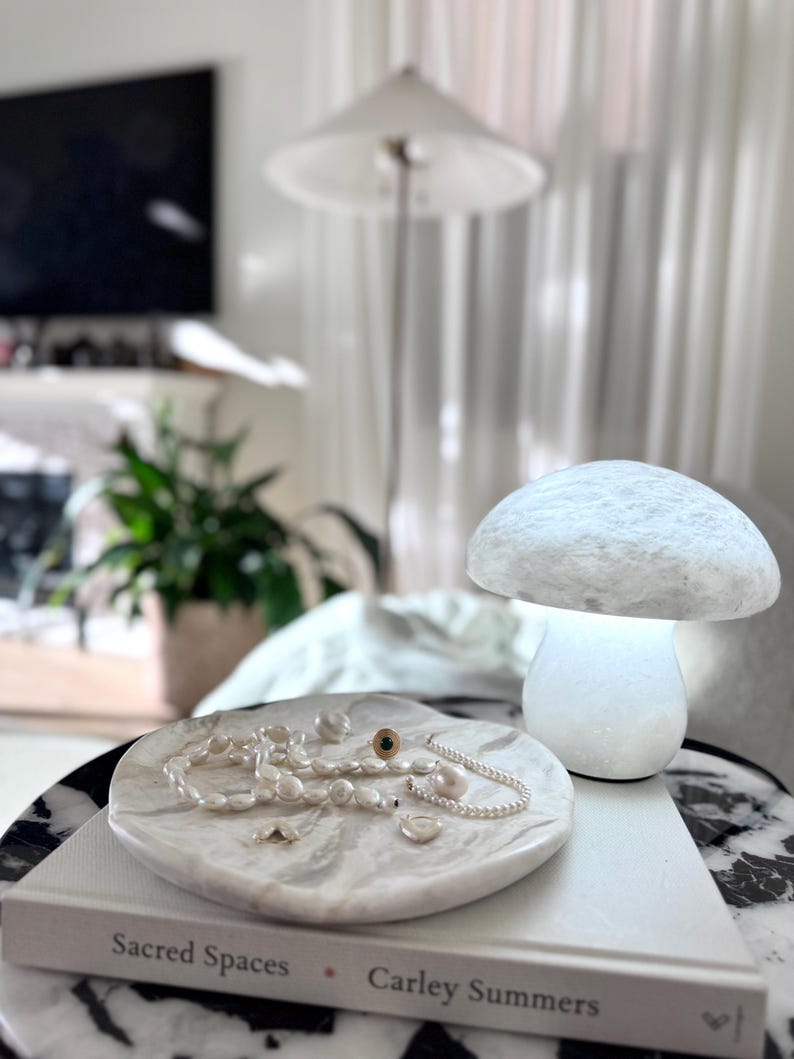 May include: A white marble tray holding a collection of pearl necklaces and a gold ring with a dark stone, placed on a book titled Sacred Spaces by Carley Summers, next to a glowing white mushroom-shaped lamp with a textured, stone-like surface.