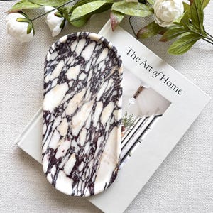 Puede incluir: Una bandeja ovalada de mármol con un patrón blanco y marrón oscuro. La bandeja está sobre un libro titulado "The Art of Home". Flores blancas y hojas verdes en el fondo.