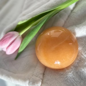 May include: An orange-colored crystal sphere rests on a white, textured surface, next to a pink tulip with green stem and leaves. The sphere has a smooth, polished appearance. The overall theme is natural and decorative.