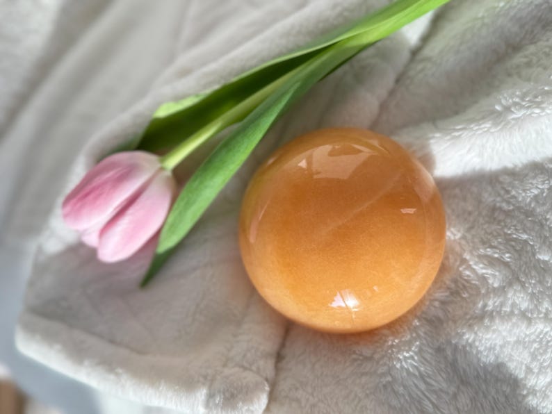 May include: An orange-colored crystal sphere rests on a white, textured surface, next to a pink tulip with green stem and leaves. The sphere has a smooth, polished appearance. The overall theme is natural and decorative.
