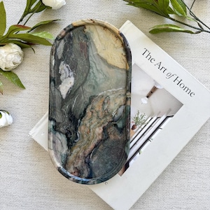May include: An oval-shaped stone tray featuring a polished surface with swirling patterns of dark green, grey, cream, and brown. The tray is positioned on top of a white book titled The Art of Home, surrounded by white flowers and green leaves.