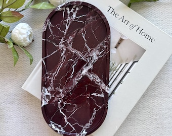 Op de afbeelding: Een ovaalvormig dienblad met een diepbordeauxrode basis en een wit gemarmerd aderpatroon. Het dienblad ligt op een wit boek met de titel The Art of Home, vergezeld door groene bladeren en kleine witte bloemen.