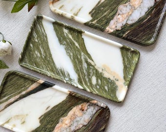 May include: Three rectangular trays featuring a marbled pattern in shades of forest green, white, and soft peach. Each tray has a smooth, polished surface with distinct veining that creates a unique, organic appearance across the set of three.