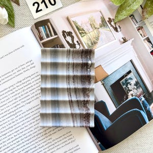 May include: A rectangular, gray and brown striped soap dish with a ridged design. The dish is resting on an open book with text and a white card with the number 210.