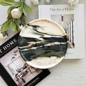 May include: A round decorative plate with a white rim and a marbled design in shades of green, black, and white. The plate is resting on a book titled "The Art of Home" and another book by Brian Paquette.
