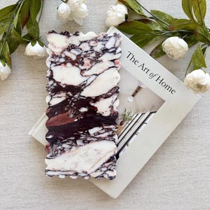 May include: A rectangular, decorative soap dish with a wavy edge, featuring a marble pattern in shades of white, burgundy, and brown. The soap dish rests on a book titled "The Art of Home" and is surrounded by white flowers and green leaves.