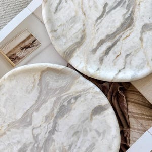 May include: Two circular trays crafted from white marble with prominent grey and beige veining. The trays feature a smooth, polished surface with natural stone patterns, showcasing swirling organic textures throughout the round stone material.