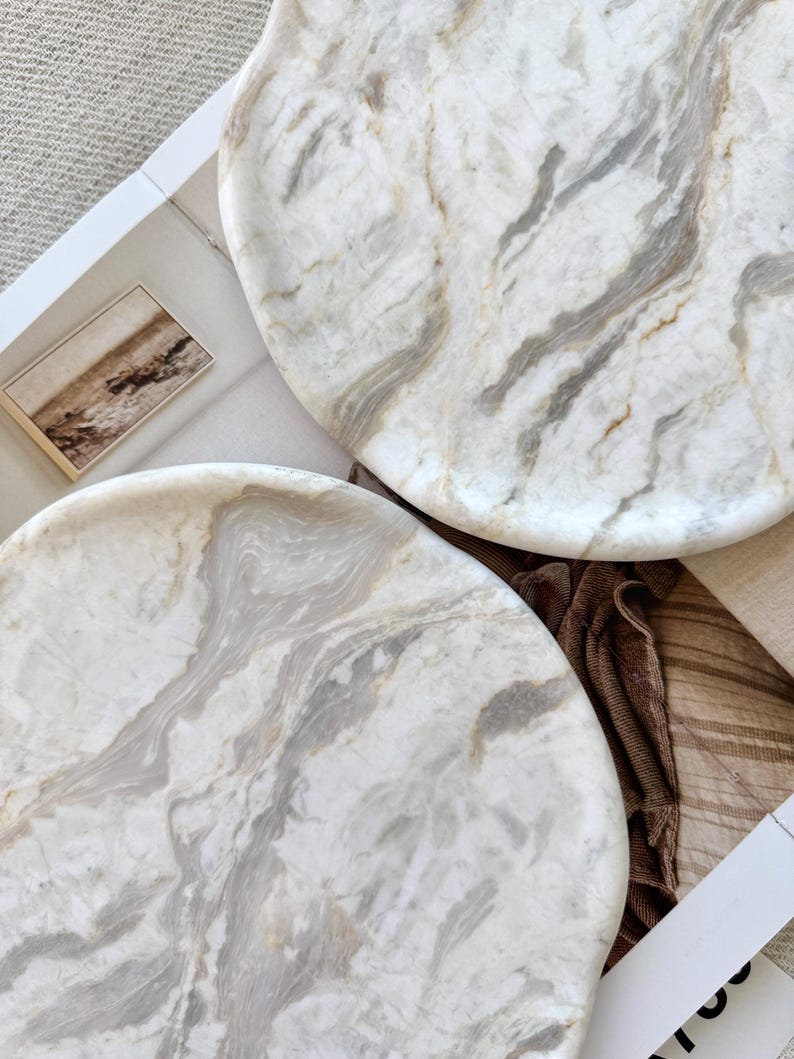 May include: Two circular trays crafted from white marble with prominent grey and beige veining. The trays feature a smooth, polished surface with natural stone patterns, showcasing swirling organic textures throughout the round stone material.