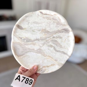 May include: A circular stone plate featuring a polished surface with swirling patterns of white, cream, and light gray veins. The edges of the plate are slightly irregular and organic in shape. A white tag with the black printed text A789 is attached.