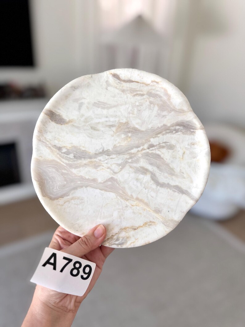 May include: A circular stone plate featuring a polished surface with swirling patterns of white, cream, and light gray veins. The edges of the plate are slightly irregular and organic in shape. A white tag with the black printed text A789 is attached.