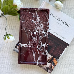May include: A rectangular tray made of deep burgundy stone with prominent white veining, resting on top of a book titled This Is Home by Natalie Walton, surrounded by several white peony flowers with green leaves on a textured fabric surface.