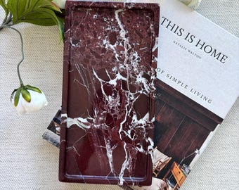 May include: A rectangular tray made of deep burgundy stone with prominent white veining, resting on top of a book titled This Is Home by Natalie Walton, surrounded by several white peony flowers with green leaves on a textured fabric surface.