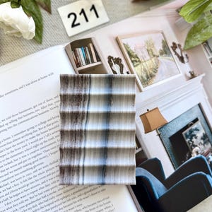 May include: A rectangular, textured coaster with a striped pattern in shades of gray, brown, and blue. The coaster is resting on an open book with text and a white background. A white number 211 is visible.