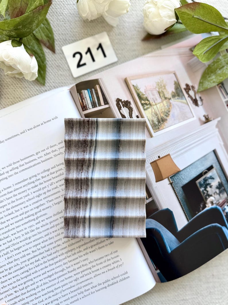 May include: A rectangular, textured coaster with a striped pattern in shades of gray, brown, and blue. The coaster is resting on an open book with text and a white background. A white number 211 is visible.