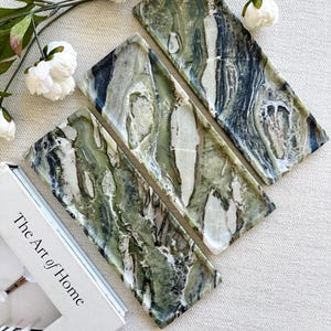 May include: Three rectangular trays crafted from polished green and white marble with distinct veining patterns. The trays are arranged diagonally next to a book with the text The Art of Home printed on the cover and white floral accents.