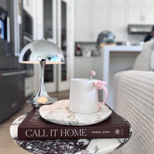 May include: A white ceramic mug with a pink bow handle and a matching pink stirrer, resting on a white marble saucer atop a dark brown book titled CALL IT HOME by Amber Lewis, positioned next to a small chrome dome lamp on a marble table.