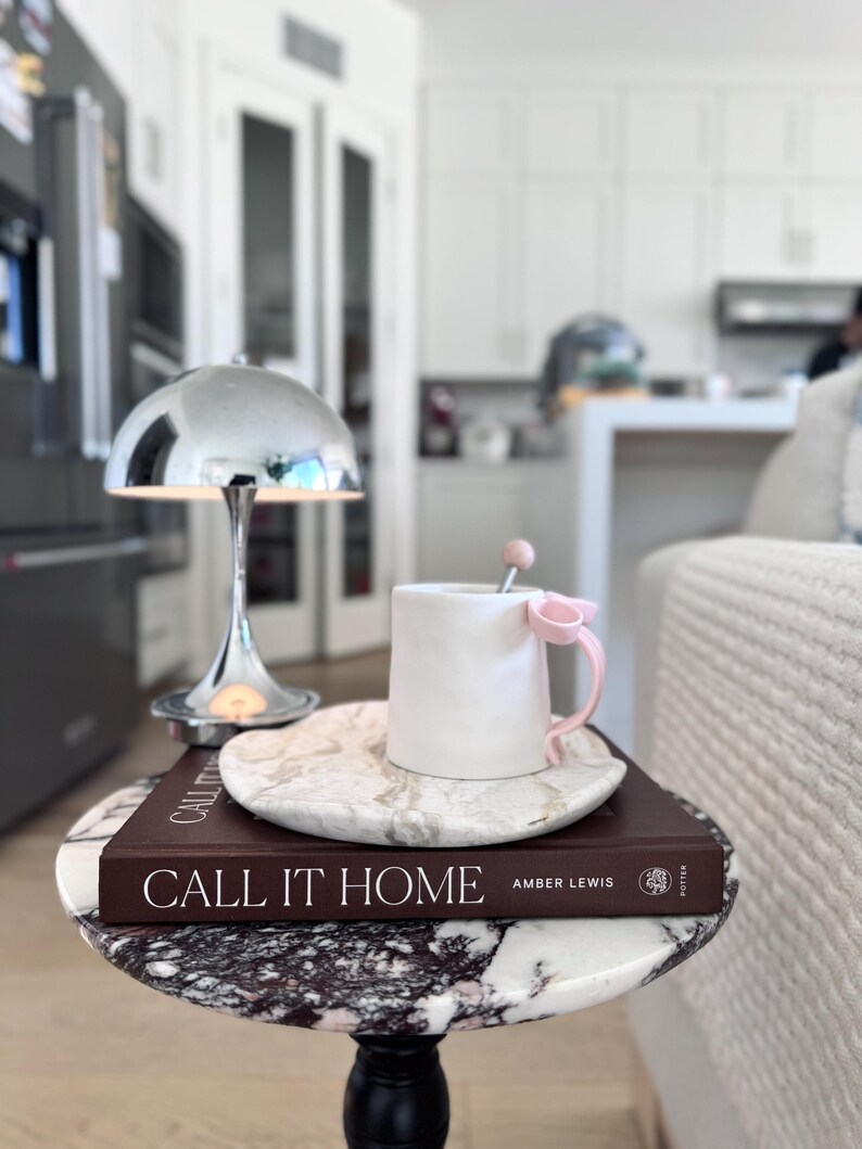 May include: A white ceramic mug with a pink bow handle and a matching pink stirrer, resting on a white marble saucer atop a dark brown book titled CALL IT HOME by Amber Lewis, positioned next to a small chrome dome lamp on a marble table.