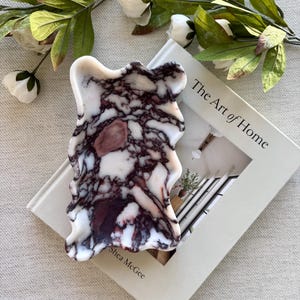 Puede incluir: Una herramienta de masaje gua sha de piedra veteada blanca y burdeos, con una forma irregular. La herramienta está colocada sobre un libro titulado "The Art of Home", con flores blancas y hojas verdes en el fondo.