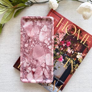 Könnte beinhalten: Ein rechteckiges Tablett mit einem rosa und dunkelburgunderfarbenen Marmormuster und einer glatten, polierten Oberfläche. Das Tablett hat erhöhte Ränder und einen flachen Boden, der ein komplexes Netzwerk aus Adern und Steinstrukturen zeigt.