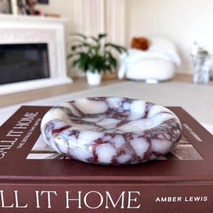 May include: A round, marble ashtray with a white base and burgundy veining. The ashtray sits on a stack of books with the title "CALL IT HOME" in white lettering. The ashtray is a decorative item for a home.