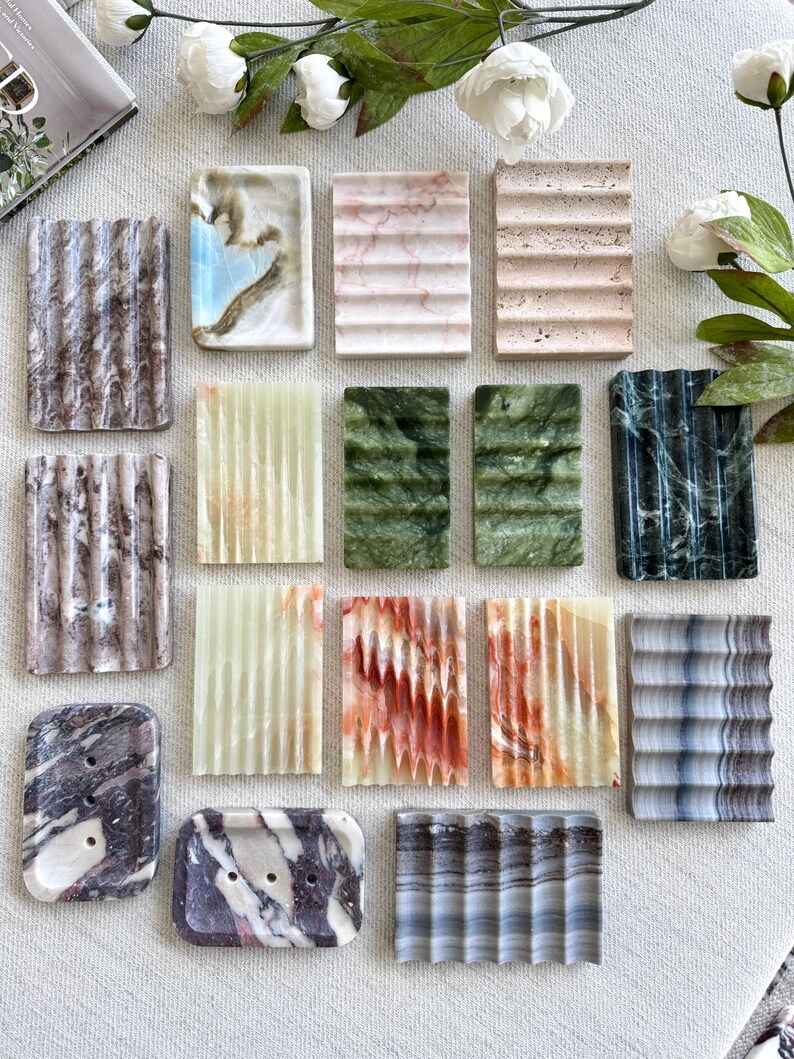 May include: A collection of rectangular soap dishes in various colors and patterns. The dishes are made of stone and feature a grooved design for drainage. Colors include white, pink, green, and gray. Some have unique veining.