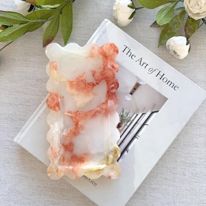 May include: A decorative, rectangular tray with a scalloped edge. The tray is white with orange and yellow marbling. The tray rests on a book titled "The Art of Home" with white flowers and greenery in the background.