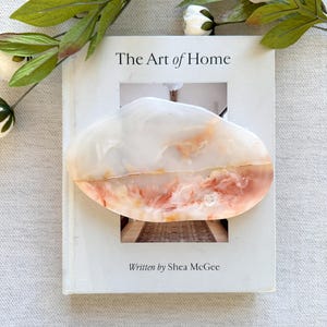 Può includere: Un oggetto decorativo ovale con un aspetto marmorizzato nei toni del bianco, pesca e marrone è appoggiato su un libro intitolato "The Art of Home" di Shea McGee. Il libro è circondato da fiori bianchi e foglie verdi.