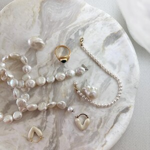 May include: A collection of jewelry including a long strand of irregular white pearls, a gold ring with a black stone, a delicate pearl bracelet, a single large baroque pearl earring, and a pair of small cream-colored heart-shaped hoop earrings.