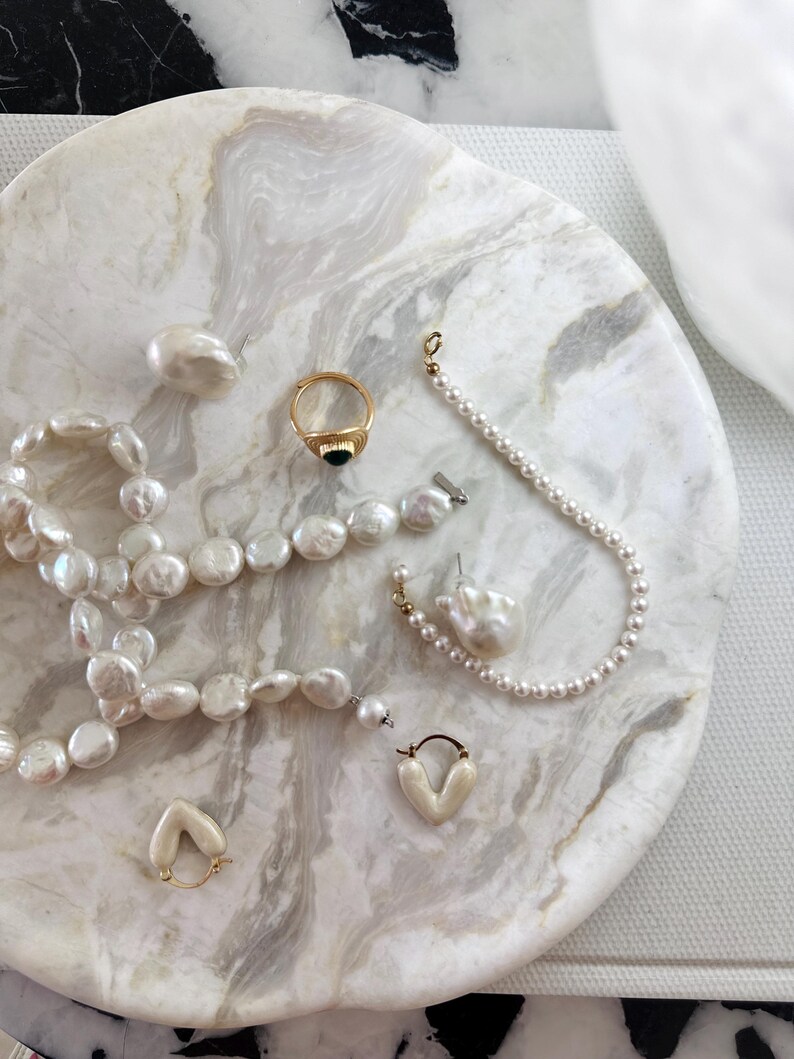 May include: A collection of jewelry including a long strand of irregular white pearls, a gold ring with a black stone, a delicate pearl bracelet, a single large baroque pearl earring, and a pair of small cream-colored heart-shaped hoop earrings.