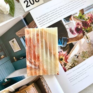May include: A rectangular, textured soap bar with a layered pattern of orange, cream, and brown hues. The soap is displayed on an open magazine with text and a white floral arrangement in the background. A white tag with the number 208 is also visible.