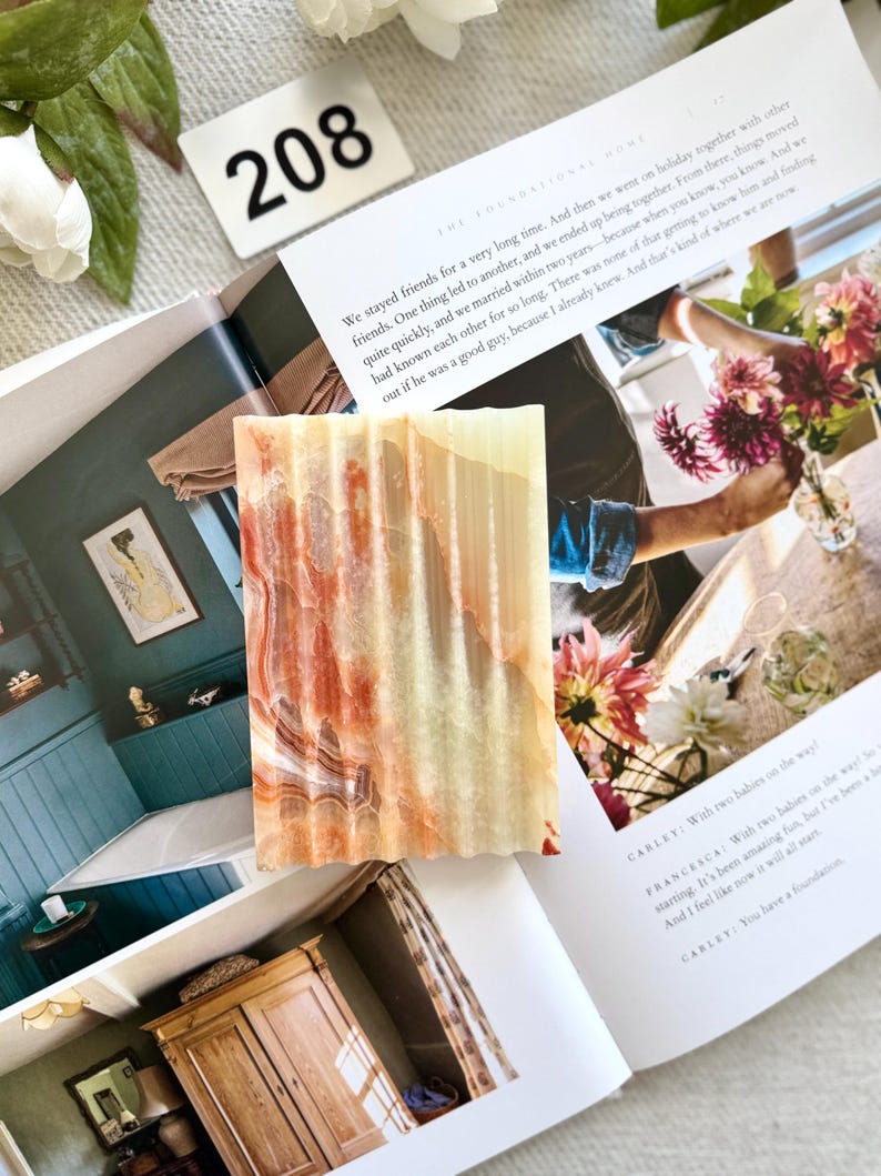 May include: A rectangular, textured soap bar with a layered pattern of orange, cream, and brown hues. The soap is displayed on an open magazine with text and a white floral arrangement in the background. A white tag with the number 208 is also visible.