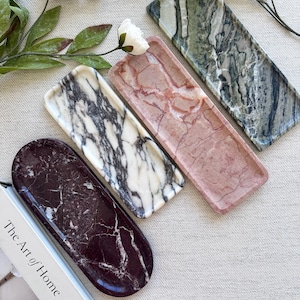 May include: Three rectangular and one oval stone tray in dark purple, white with black veining, pink, and green marble patterns. The trays are arranged diagonally next to a book with the text The Art of Home visible on the white cover.