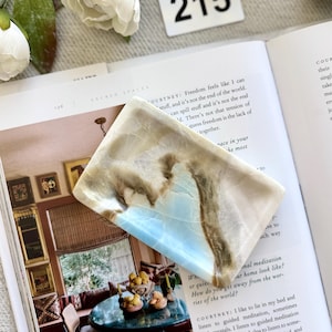 May include: A rectangular, polished stone with a light blue and brown marbled pattern. The stone is resting on an open book with the number "215" visible in the background. The book's pages display text about meditation and self-care.